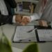 Two people in formal attire shake hands across a desk with documents, a clipboard, and a pen, suggesting an Installment Loans agreement or contract signing. A green plant is visible in the foreground. | MONEY6X