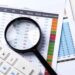 A magnifying glass and calculator placed on financial documents with charts and tables.