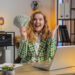 A smiling woman sitting at a desk with a laptop holds a fan of dollar bills in one hand and raises her other hand in excitement, suggesting financial success or a windfall in a modern office setting. | MONEY6X