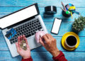 A person holds coins and a credit card over a laptop, while using the other hand to put a coin into a pink piggy bank. Nearby are a smartphone, potted plant, coffee cup, and stationery on a blue wooden table. | MONEY6X