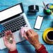 A person holds coins and a credit card over a laptop, while using the other hand to put a coin into a pink piggy bank. Nearby are a smartphone, potted plant, coffee cup, and stationery on a blue wooden table. | MONEY6X
