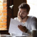 A man sits at a table with a laptop and coffee cup, holding a piece of paper and looking concerned or stressed, resting his face on his hand in a brightly lit room with brick walls and plants. | MONEY6X