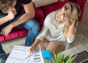 A woman and a man sit at a glass table with papers, a blue calculator, and a laptop, looking concerned as they review documents together in a living room with a red sofa. | MONEY6X