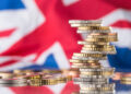Stacks of British coins are placed in front of a blurred Union Jack flag, symbolizing the United Kingdom’s economy or financial matters. | MONEY6X