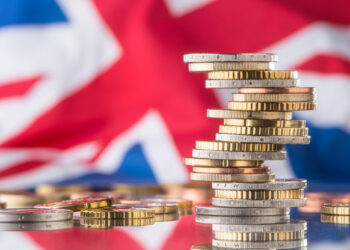 Stacks of British coins are placed in front of a blurred Union Jack flag, symbolizing the United Kingdom’s economy or financial matters. | MONEY6X
