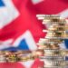 Stacks of British coins are placed in front of a blurred Union Jack flag, symbolizing the United Kingdom’s economy or financial matters. | MONEY6X