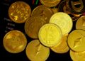 A pile of shiny gold-colored cryptocurrency coins, including Bitcoin, Ethereum, and Dogecoin, displayed on a dark background with faint financial chart lines. | MONEY6X