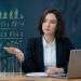 A businesswoman in a suit sits at a desk with a laptop, notebook, and books, presenting virtual financial graphs and charts with her hand, in a modern office setting. | MONEY6X