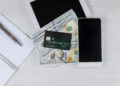 A smartphone, a tablet, a notebook with a pen, and a credit card placed on top of several hundred-dollar bills are arranged on a light wooden surface. | MONEY6X