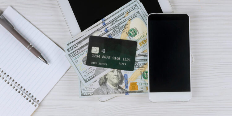 A smartphone, a tablet, a notebook with a pen, and a credit card placed on top of several hundred-dollar bills are arranged on a light wooden surface. | MONEY6X