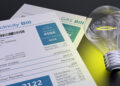 A close-up of electricity and gas bills next to a glowing light bulb on a dark surface, symbolizing energy usage and costs. The electricity bill shows the total amount due as 450 euros. | MONEY6X