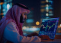A man in traditional Middle Eastern attire analyzes colorful financial charts on a laptop at night, with blurred city lights in the background. | MONEY6X