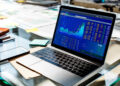 A laptop displaying colorful stock market charts and financial data sits on a glass desk, surrounded by papers, sticky notes, and a smartphone. | MONEY6X