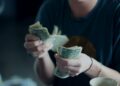 A person wearing a dark shirt is counting a stack of U.S. one-dollar bills, with a coffee cup visible in the foreground. Their face is out of focus and not fully visible. | MONEY6X