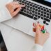 Person in a white sweater holding a credit card and typing on a laptop keyboard, possibly shopping or making an online payment. | MONEY6X