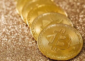 Five gold Bitcoin coins placed in a line on a shimmering, glittery golden surface. | MONEY6X