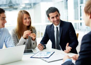 A group of four professionals, three men and one woman, are engaged in a discussion in a modern office. Dressed in business attire and surrounded by documents, a laptop, and a tablet on the table before them, they seem to be evaluating how many jobs are available in real estate investment trusts. | MONEY6X