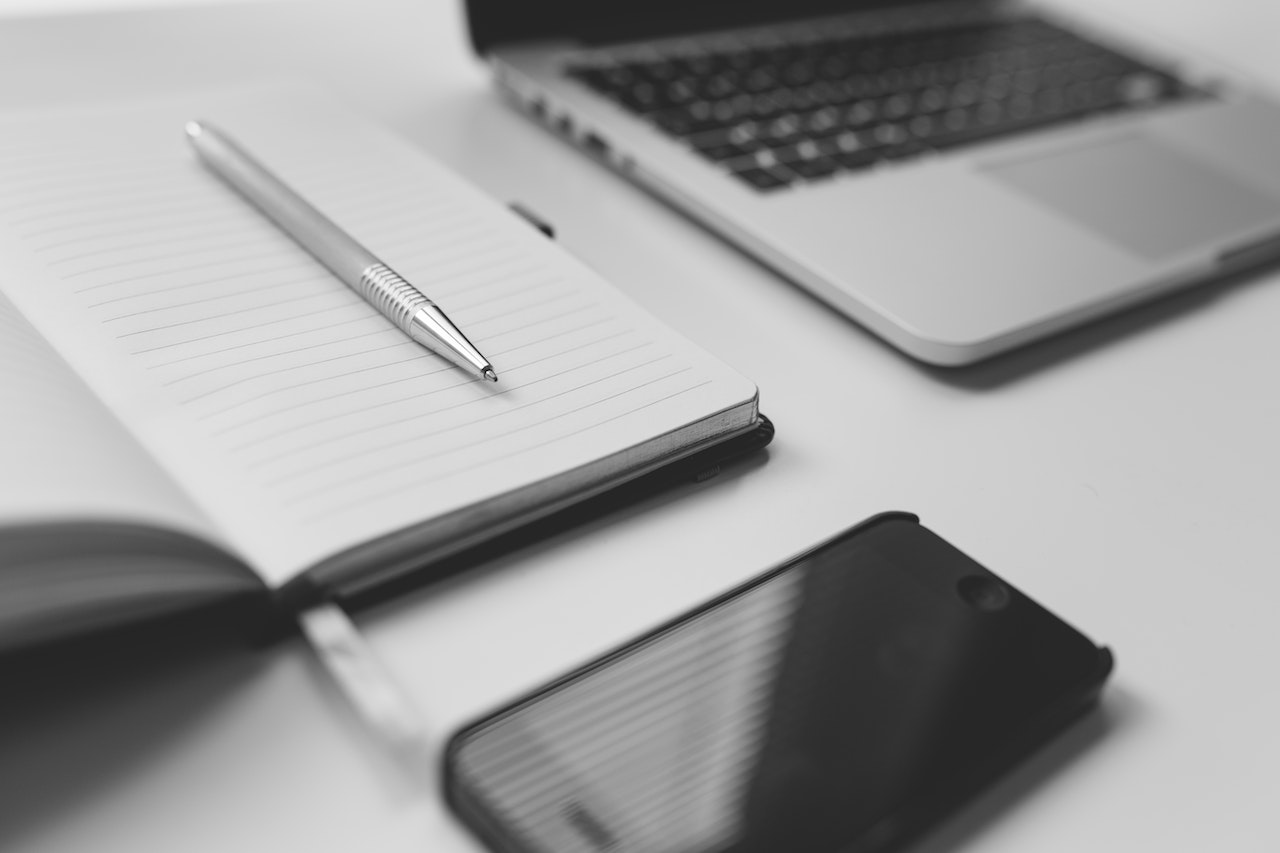 A black and white image of an open notebook with lined pages and a pen placed horizontally on top. Next to the notebook, there is a partially visible laptop and a smartphone lying face up, perhaps ready for remote real estate jobs. The scene appears to be set on a clean, white desk. | MONEY6X