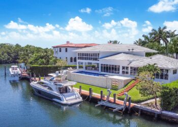 A luxurious waterfront property featuring a large modern house with a white exterior, expansive windows, and a spacious outdoor pool. This best real estate investment offers a private dock with boats moored, set against lush greenery and a serene waterway under a bright blue sky. | MONEY6X