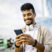 A smiling person with curly hair and wearing headphones around their neck is holding and looking at a smartphone, possibly exploring apps to make money. They are dressed in a beige jacket over a white shirt, standing outdoors with a blurred background of buildings and a blue sky. | MONEY6X
