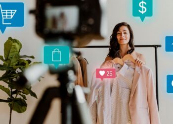 A woman holding clothes on hangers stands in front of a camera on a tripod, surrounded by social media and e-commerce icons like a shopping cart, dollar sign, heart with 25 likes, house, and chat bubble with 9 messages. A plant is visible in the background as she shares tips to make money fast. | MONEY6X