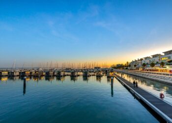 A serene harbor at sunset with numerous docked boats reflecting on the calm water in Lisbon, Portugal. A wooden pier extends into the water, and modern buildings line the waterfront on the right side. The sky transitions from blue to a warm golden hue, showcasing prime real estate opportunities. | MONEY6X