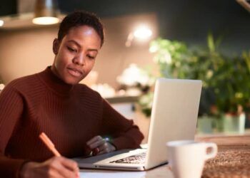 A person with short hair is sitting at a table in a cozy, dimly-lit room, using a laptop and writing notes on paper. They are wearing a brown sweater, and a coffee mug is placed on the table beside them. There are blurred plants in the background as they focus on applying the 50/30/20 Rule. | MONEY6X