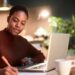 A person with short hair is sitting at a table in a cozy, dimly-lit room, using a laptop and writing notes on paper. They are wearing a brown sweater, and a coffee mug is placed on the table beside them. There are blurred plants in the background as they focus on applying the 50/30/20 Rule. | MONEY6X