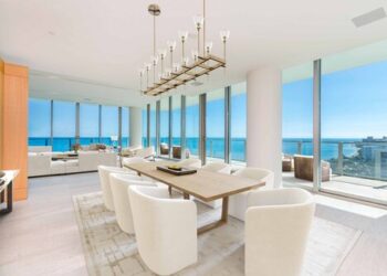 A modern dining area in an upscale apartment features a wooden dining table surrounded by white chairs. A geometric chandelier hangs above. Floor-to-ceiling windows, virtually staged by top companies, offer a stunning ocean view with clear blue skies and sunlight illuminating the room. | MONEY6X