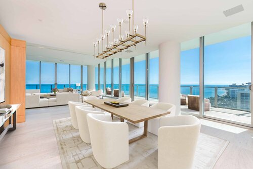 A modern dining area in an upscale apartment features a wooden dining table surrounded by white chairs. A geometric chandelier hangs above. Floor-to-ceiling windows, virtually staged by top companies, offer a stunning ocean view with clear blue skies and sunlight illuminating the room. | MONEY6X