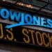 The image shows an electronic ticker sign displaying "DOW JONES" in blue and "U.S. STOCKS" in yellow lights underneath, reflecting stock market information. The background includes part of a building with a dark facade, reminiscent of Dow Jones Fintechzoom updates. | MONEY6X