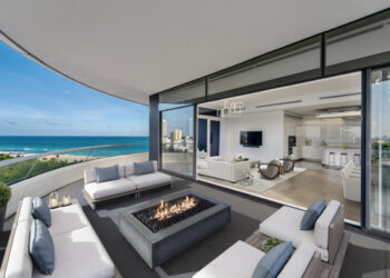 A modern oceanfront apartment balcony with a cozy seating area around a square fire pit. Glass doors lead into a sleek living area featuring contemporary furnishings, a large TV, and an open kitchen. Enhanced by virtual home staging, the balcony offers stunning ocean and city views. | MONEY6X