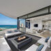 A modern oceanfront apartment balcony with a cozy seating area around a square fire pit. Glass doors lead into a sleek living area featuring contemporary furnishings, a large TV, and an open kitchen. Enhanced by virtual home staging, the balcony offers stunning ocean and city views. | MONEY6X