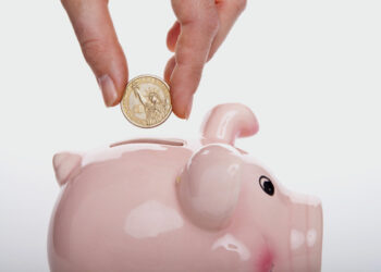 A frugal hand is shown inserting a coin into a pink piggy bank. The coin features an image resembling the Statue of Liberty. The background is plain white, emphasizing the piggy bank and the hand holding the coin. | MONEY6X