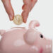 A frugal hand is shown inserting a coin into a pink piggy bank. The coin features an image resembling the Statue of Liberty. The background is plain white, emphasizing the piggy bank and the hand holding the coin. | MONEY6X