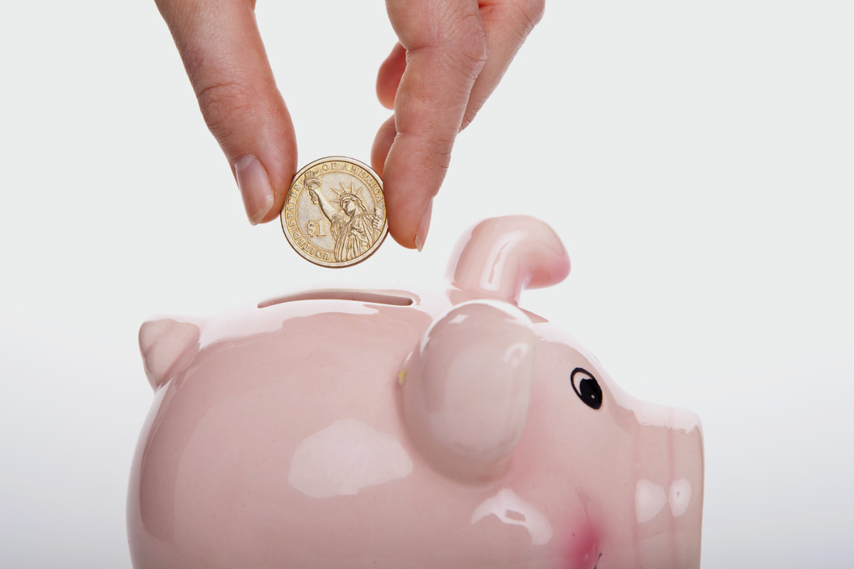 A frugal hand is shown inserting a coin into a pink piggy bank. The coin features an image resembling the Statue of Liberty. The background is plain white, emphasizing the piggy bank and the hand holding the coin. | MONEY6X