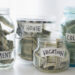 Four glass jars filled with U.S. dollar bills are labeled “Retirement,” “House,” “Vacation,” and “College.” The labels, written on pieces of paper taped to the jars, signify savings goals. These jars, arranged in a line against a light background, illustrate some of the best ways to save money effectively. | MONEY6X