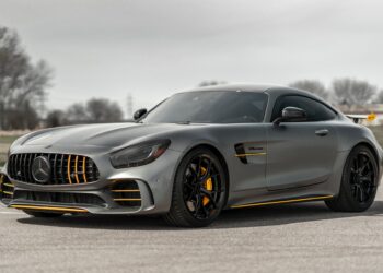 A sleek, grey luxury sports car with yellow accents is parked on an empty road. The car has a low profile, aerodynamic design, and black alloy wheels. The background features a blurred landscape with bare trees and a cloudy sky. Imagine cruising in style while you also save money on car insurance. | MONEY6X