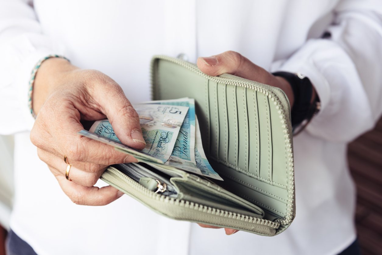 A person wearing a white shirt is holding an open green wallet and handling some British pound notes, probably thinking, "I need money now." Several card slots in the wallet are visible, but no cards are present. The wallet's zipper and compartments are also clearly visible. | MONEY6X