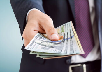 A person in a dark suit and tie extends their hand, holding a stack of US 100-dollar bills. The background is blurred with a light blue gradient, focusing attention on the money, hinting at an opportunity to make 6x more money. | MONEY6X