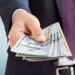 A person in a dark suit and tie extends their hand, holding a stack of US 100-dollar bills. The background is blurred with a light blue gradient, focusing attention on the money, hinting at an opportunity to make 6x more money. | MONEY6X