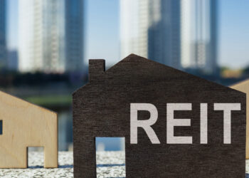 A wooden house-shaped cutout with the word "REIT" on it is placed in the foreground, while two smaller wooden house cutouts are nearby. Tall, blurry skyscrapers stand in the background, suggesting an urban setting. The image symbolizes real estate investment and highlights dividend history as a key feature. | MONEY6X