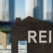 A wooden house-shaped cutout with the word "REIT" on it is placed in the foreground, while two smaller wooden house cutouts are nearby. Tall, blurry skyscrapers stand in the background, suggesting an urban setting. The image symbolizes real estate investment and highlights dividend history as a key feature. | MONEY6X