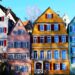 A row of colorful, traditional European houses stands under a clear blue sky. The buildings, perfect for Germany for real estate investment, are painted in shades of blue, pink, yellow, and green with contrasting window shutters. Trees and shrubs can be seen in the foreground. | MONEY6X