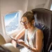 A woman with long hair tied in a high ponytail, wearing glasses and a sleeveless top, sits in an airplane seat by the window, reading an article titled "How to Save Money on Flights" on her smartphone with an airplane wing and clouds visible through the window. | MONEY6X