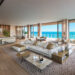 A spacious, modern living room with beige and neutral tones features plush seating, large windows offering a stunning ocean view, wooden flooring, a contemporary kitchen with a wine display, and a stylish dining area. Considering selling your home quickly? This space opens to a balcony with outdoor seating. | MONEY6X