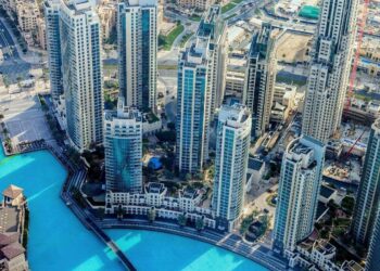 An aerial view of a modern cityscape with high-rise buildings surrounding a large blue pool. The area features contemporary architecture, lush greenery, and well-planned infrastructure. Several roads and smaller structures can be seen in the background—an ideal scene to invest in Dubai's real estate. | MONEY6X