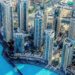 An aerial view of a modern cityscape with high-rise buildings surrounding a large blue pool. The area features contemporary architecture, lush greenery, and well-planned infrastructure. Several roads and smaller structures can be seen in the background—an ideal scene to invest in Dubai's real estate. | MONEY6X