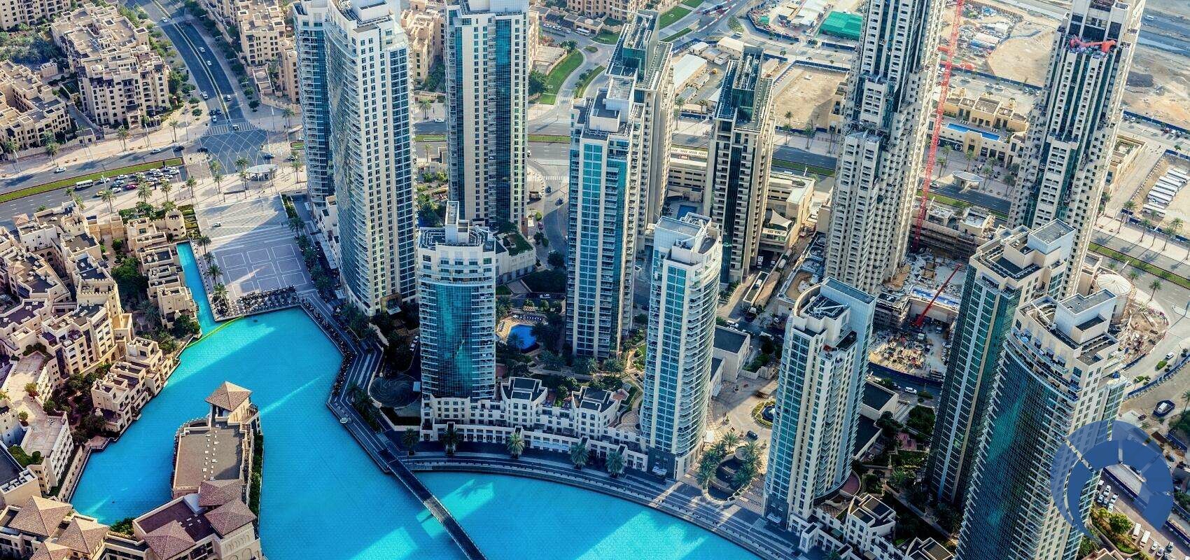 An aerial view of a modern cityscape with high-rise buildings surrounding a large blue pool. The area features contemporary architecture, lush greenery, and well-planned infrastructure. Several roads and smaller structures can be seen in the background—an ideal scene to invest in Dubai's real estate. | MONEY6X