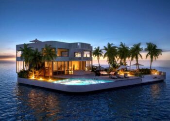 A luxurious floating home is surrounded by calm ocean waters at sunset. The modern structure features large windows, a swimming pool with loungers, and is adorned with vibrant lighting. Tropical palm trees line the house, adding a serene and exotic atmosphere. | MONEY6X